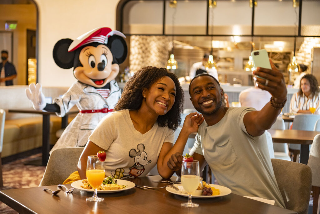 Conocer personajes en Disney World en restaurantes seleccionados. Una pareja comiendo mientras Minnie Mouse posa para una foto al fondo